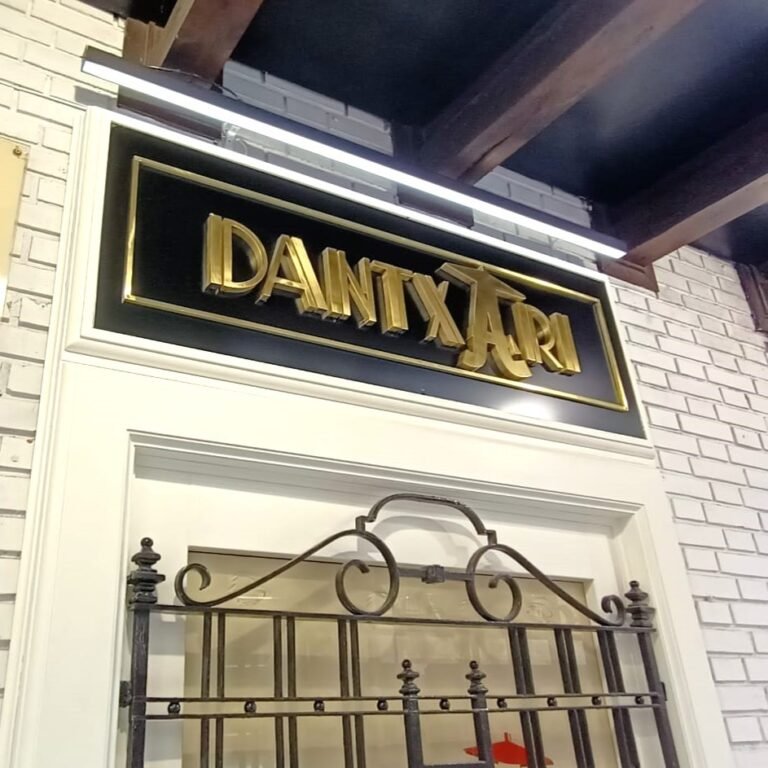 Rótulo con letras corpóreas de latón dorado brillante sobre fondo negro, montado en fachada interior con moldura blanca y reja decorativa de forja